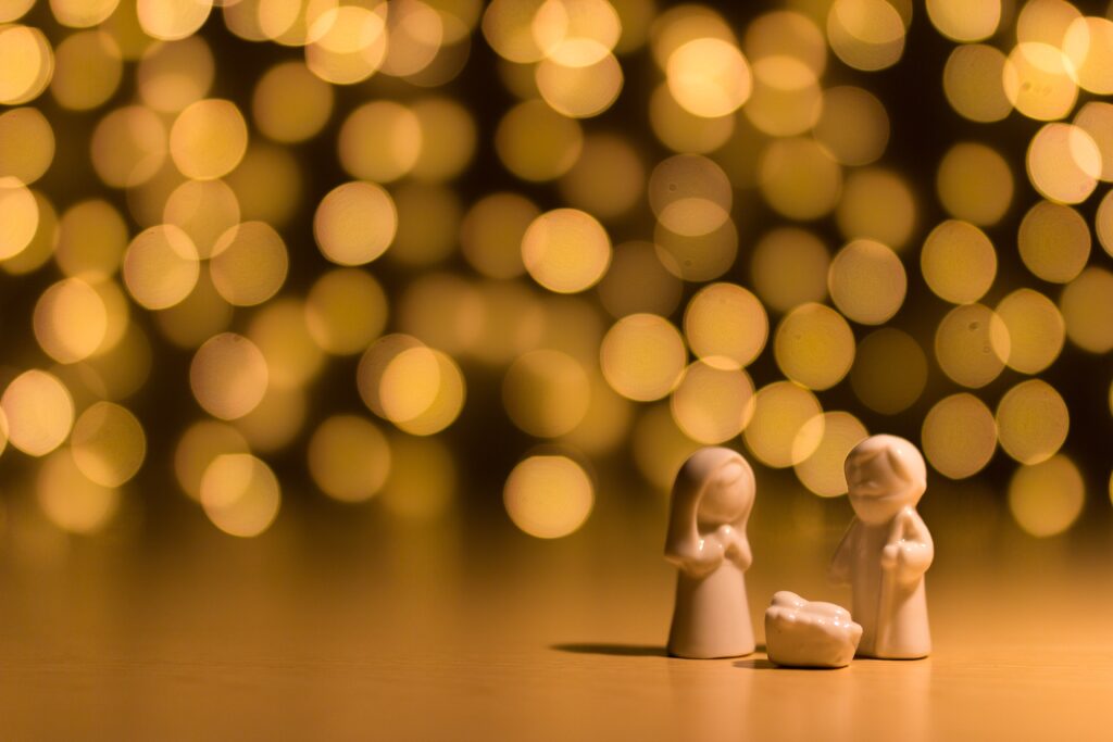 picture of Mary, Joseph and baby Jesus in a manger carvings in off white on a plain table with glitter lights behind