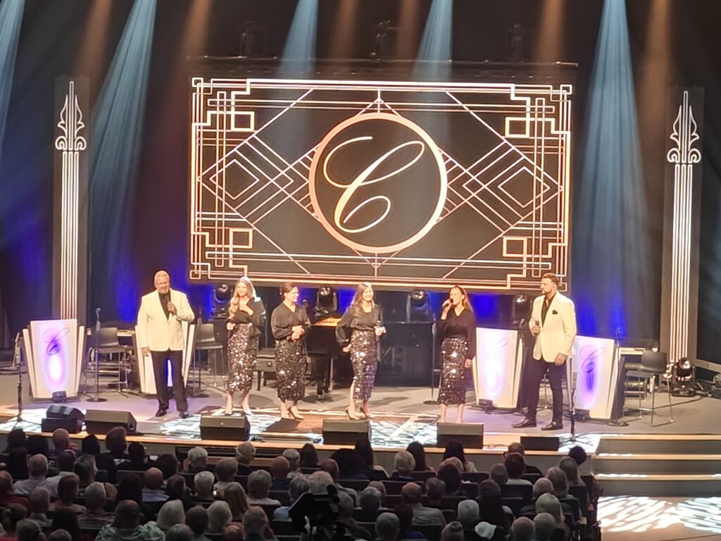 Collingsworth family on stage with lights, a backdrop with a large C. The four ladies are wearing black blouses and black sparkly skirts. The two men ad in white suit jackets with black pants and black shirts.