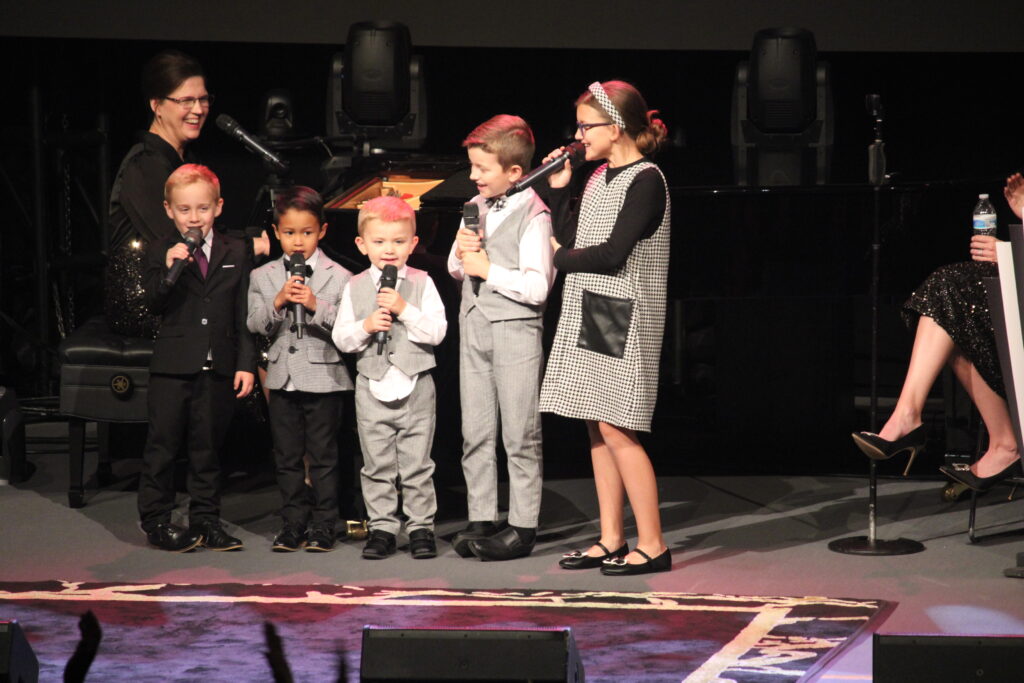 five Collingsworth grandchildren, four boys and a girl ranging in age from 3-9 standing on stage singing into microphones as Grandmother, Kim, plays the piano for them.