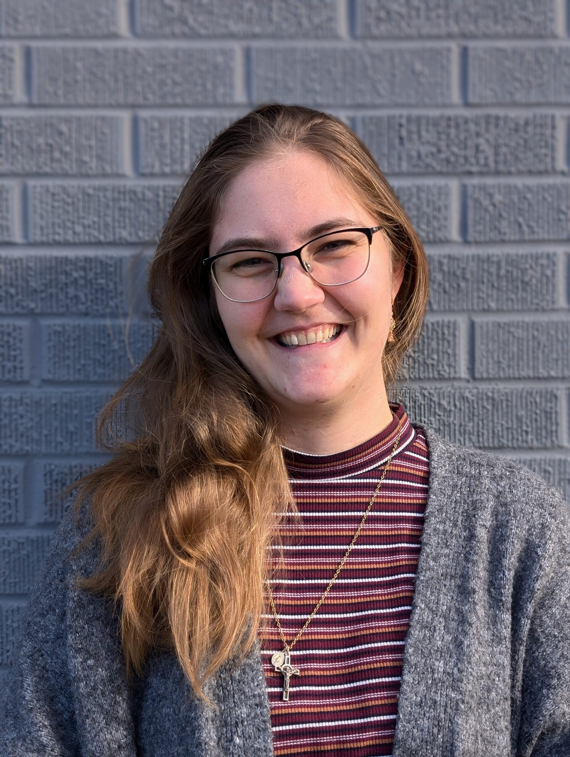 a headshot of a smiling woman with glasses and long light brown hair wearing a burgunday and grey striped shirt with a grey sweater and long silver necklace