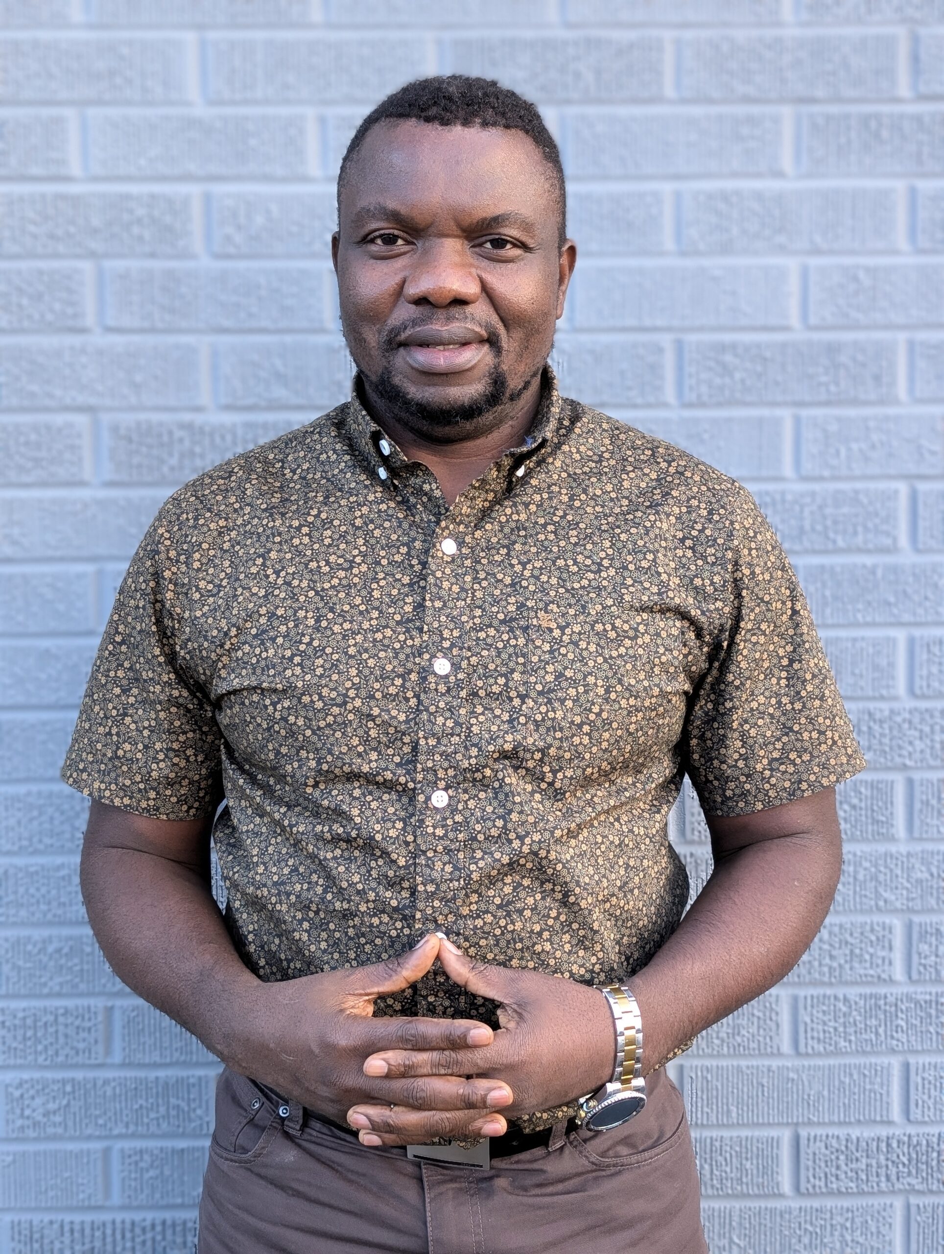 a headshot of a smiling man with dark skin and dark hair wearing a black/tan patterned shirt