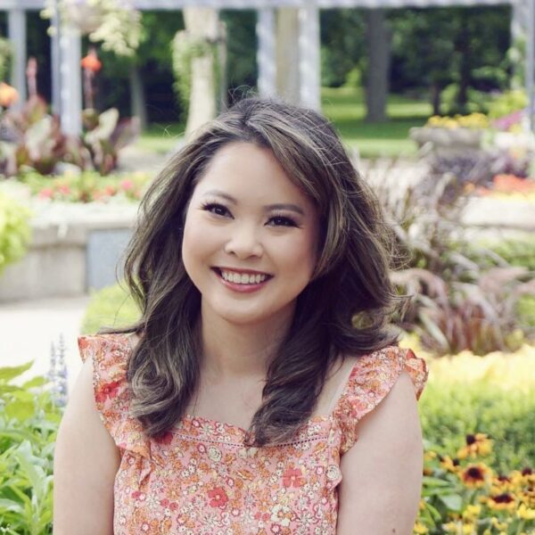 Photo of a young Vietnamese woman with long hair, wearing a short sleeved peach coloured summer dress, while sitting in a garden