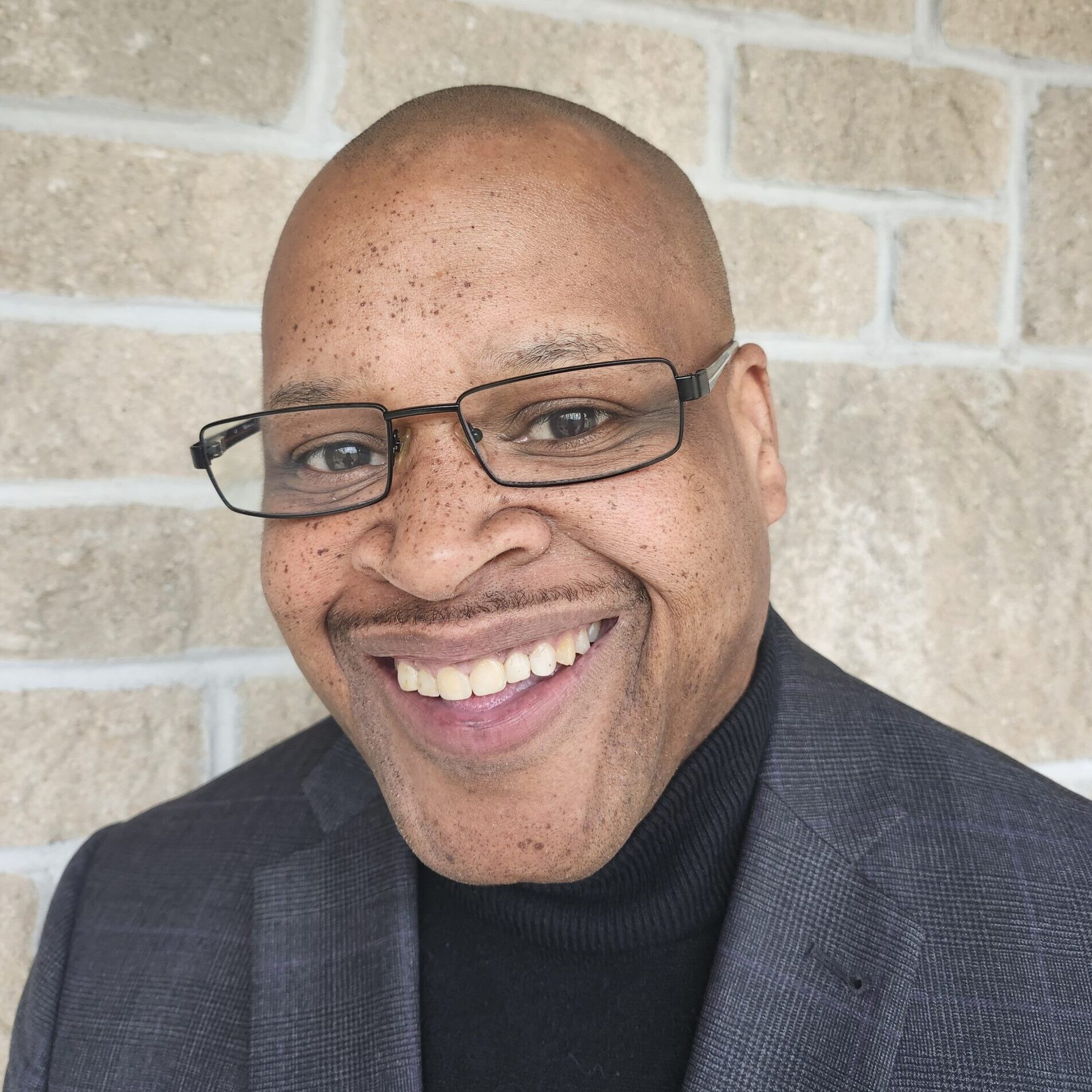 a headshot of a smiling bald man with dark skin wearing glasses, a grey suit jacket and a black turtleneck sweater