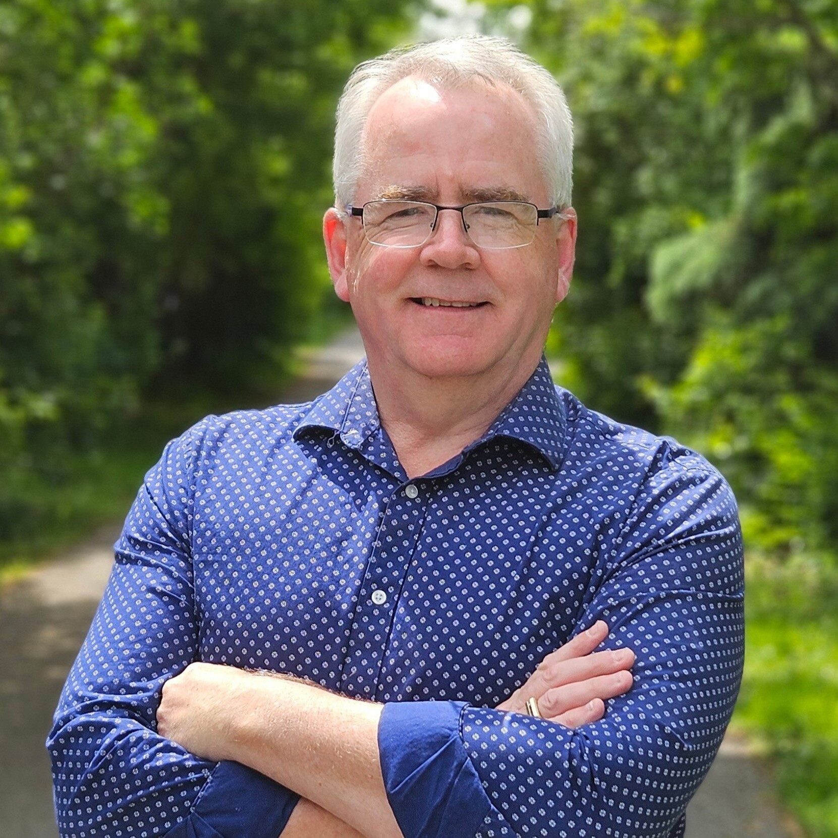 a headshot of a smiling man with white hair and glasses, wearing a blue dress shirt with arms crossed in front of him