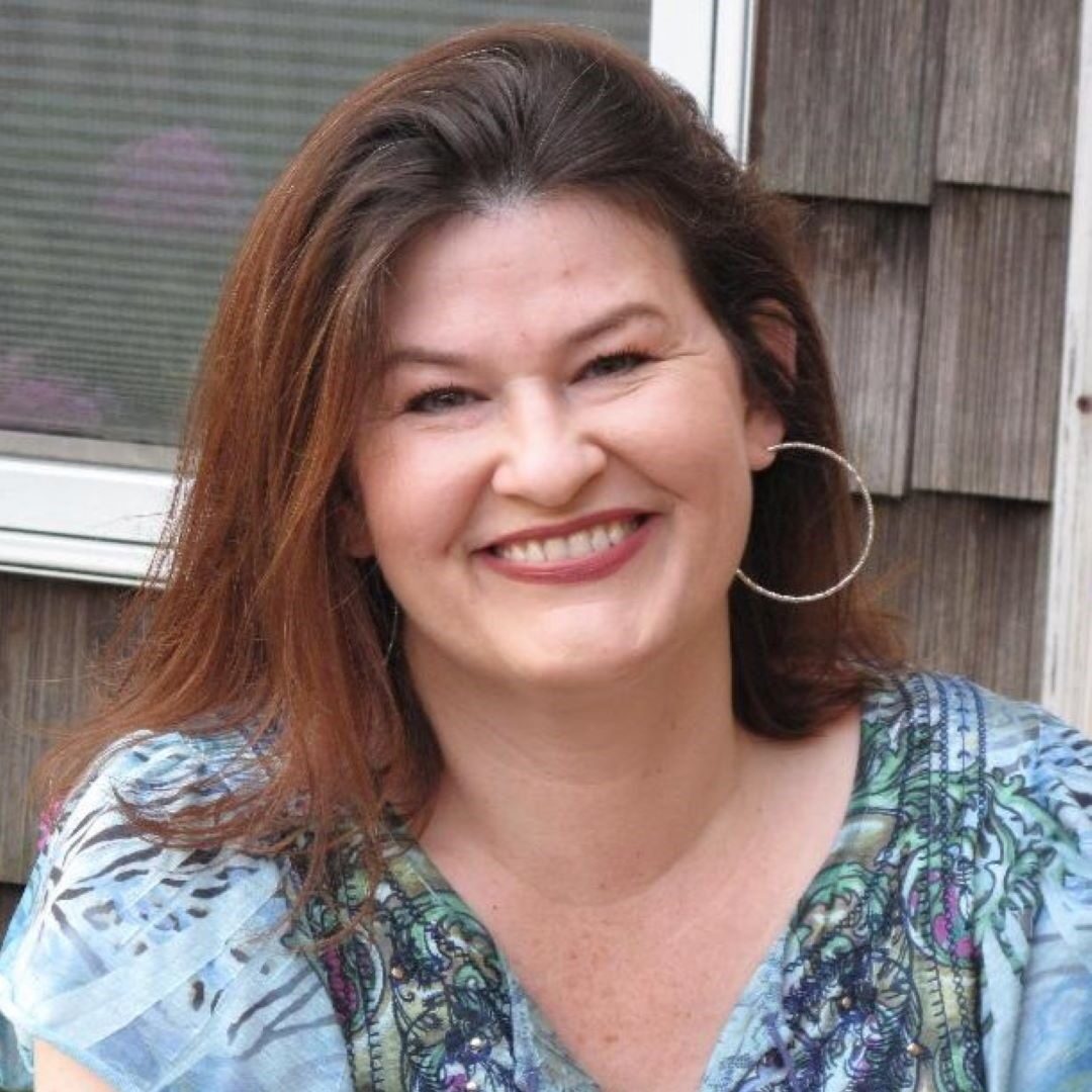 a headshot of a smiling woman with shoulder length brown hair with hoop earrings and a blue/green patterned top