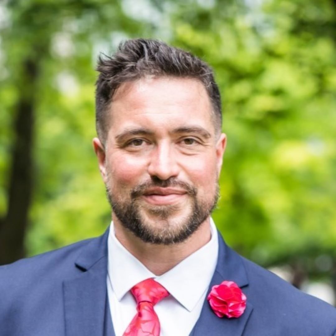 a headshot of a man with dark hair wearing a blue suit, white shirt with red tie and red carnation on his lapel