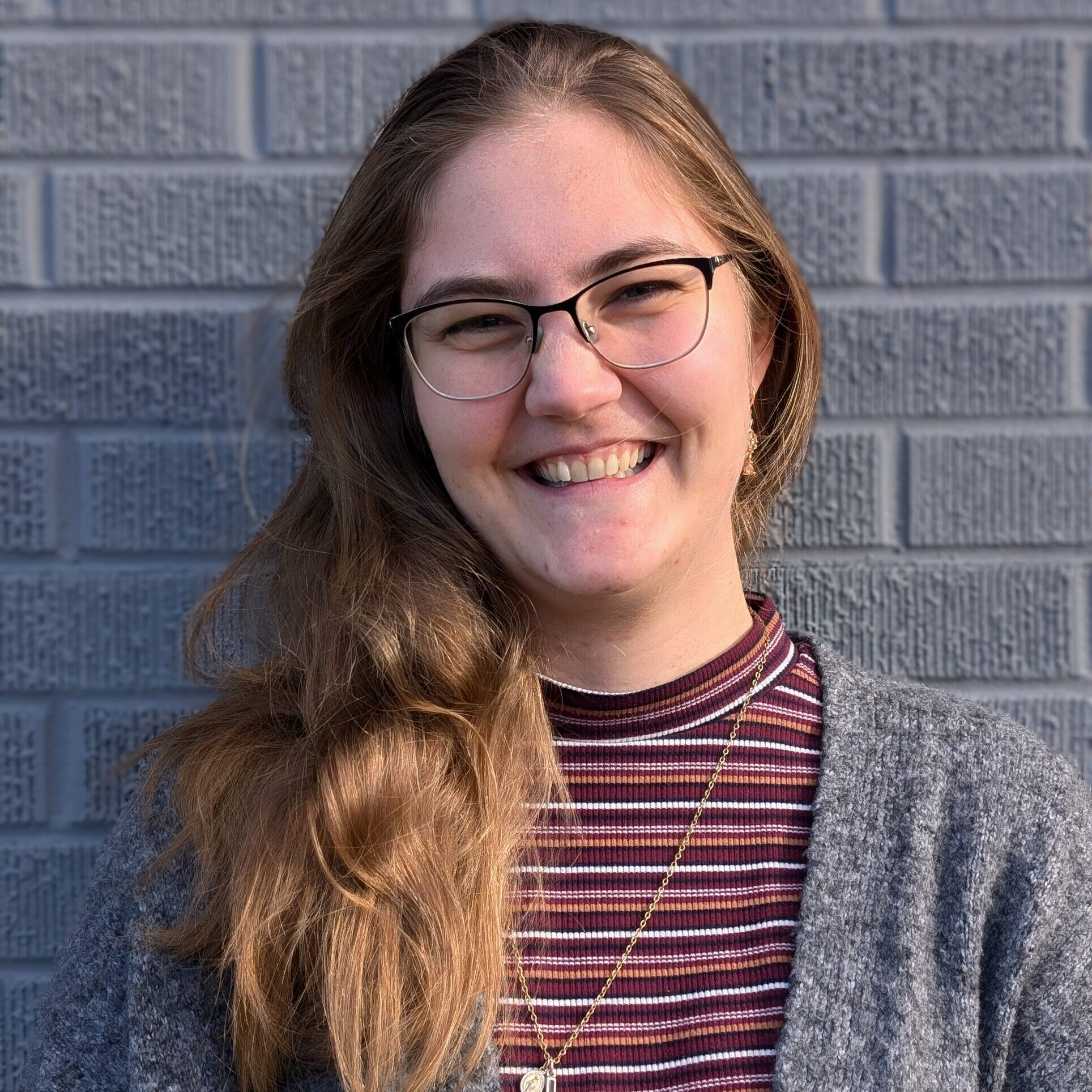 a headshot of a smiling woman with glasses and long light brown hair wearing a burgunday and grey striped shirt with a grey sweater and long silver necklace