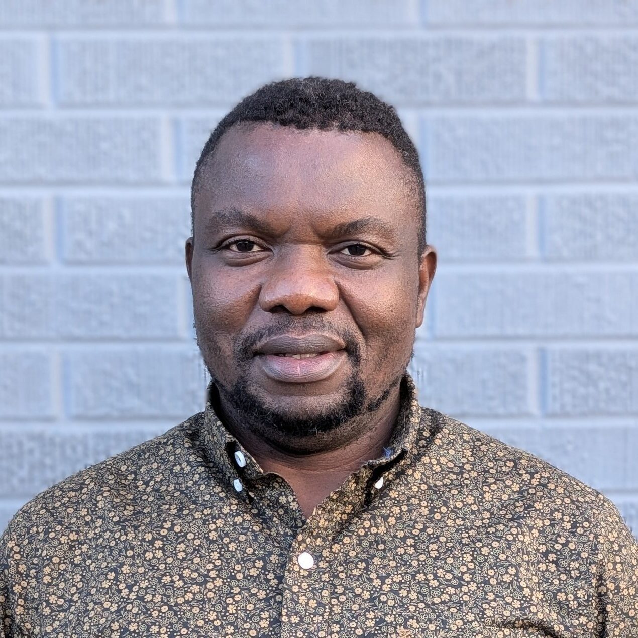 a headshot of a smiling man with dark skin and dark hair wearing a black/tan patterned shirt