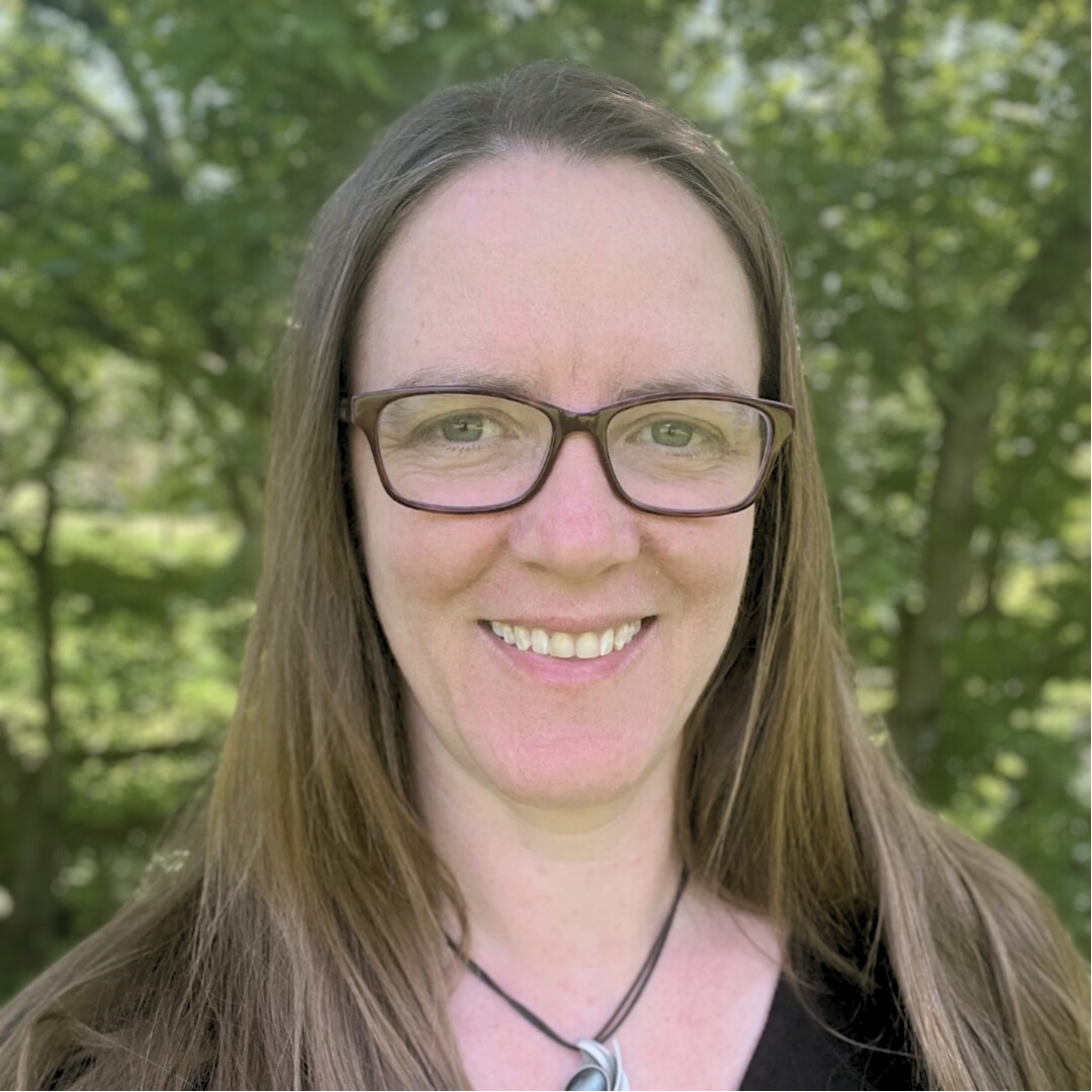 a headshot of a smiling woman with glasses, long brown hair, wearing a necklace and black top