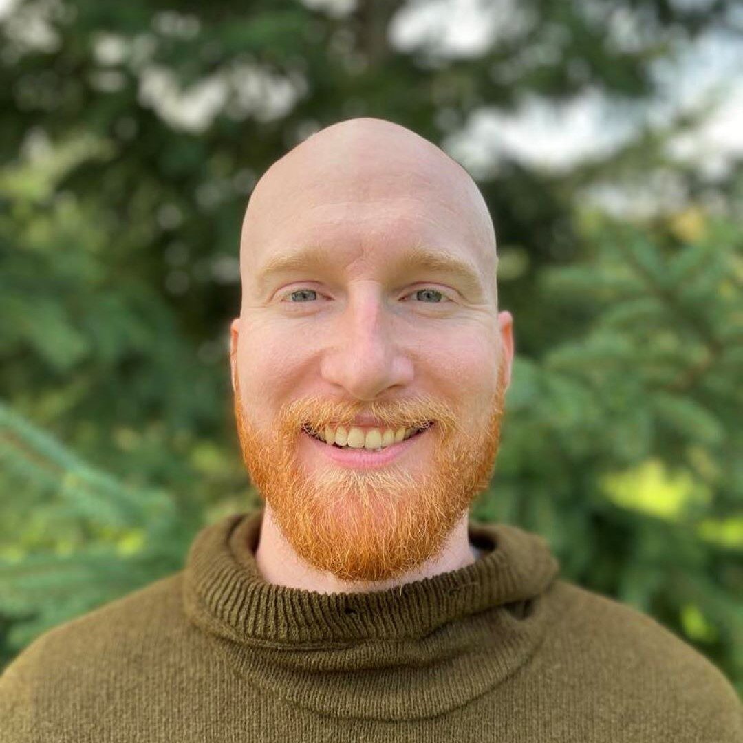 a headshot of a smiling bald man with a red beard and moustache wearing a olive green turtleneck sweater